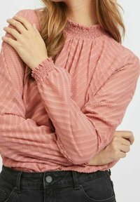 Blusa rosa con strisce testurizzate, colletto arricciato e maniche lunghe, abbinata a un denim scuro. Tessuto morbido e arricciato.