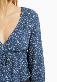 Top floreale blu con maniche lunghe, scollo a V profondo e vita arricciata con un nastro. Realizzato in tessuto leggero con piccoli motivi di fiori bianchi.