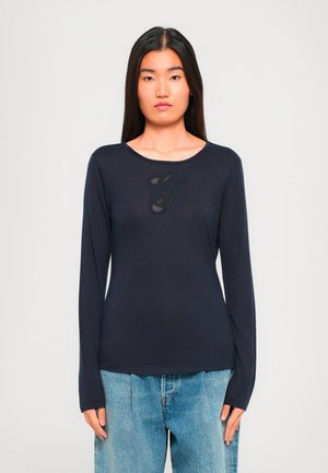 Donna con lunghi capelli scuri che indossa una maglietta nera a maniche lunghe con un design sottile e jeans azzurri larghi, in piedi contro uno sfondo bianco.