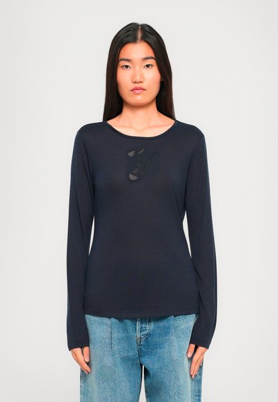 Femme aux longs cheveux bruns portant un t-shirt noir à manches longues avec un design subtil et un jean léger bleu clair, se tenant devant un fond blanc.