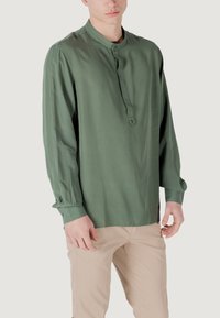 Camicia a maniche lunghe in tessuto verde con scollatura rotonda, chiusura frontale con bottoni e dettagli plissettati sulle maniche. Indossata con pantaloni beige.