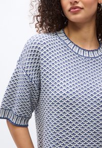 Γυναίκα που φοράει μπλε και λευκό πλεκτό πουλόβερ με κοντά μανίκια και ριμπ γιακά και μανσέτες, με κατσαρά μαλλιά και μικρούς κρίκους σκουλαρίκια.