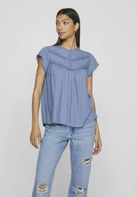 Blusa azzurro chiaro a maniche corte con vita arricciata, dettagli in pizzo sul corpetto e scollatura rotonda. Indossata con jeans in denim strappati.