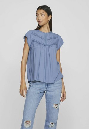 Light blue short-sleeve blouse with gathered waist, lace detailing on bodice, and rounded neckline. Worn with distressed denim jeans.