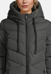 Frau trägt eine dunkelgraue gepolsterte Winterjacke mit hohem Kragen, Kapuze, schwarzem Reißverschluss und schwarzen Kordeln.