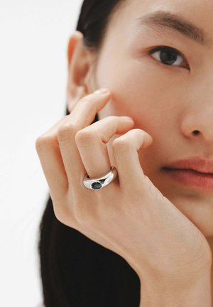 Anillo de plata con un acabado liso y pulido y una piedra azul oscura engastada en una banda curva, llevado en una mano con un tono de piel natural y texturizado.