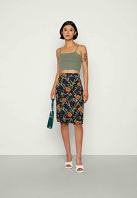 Mulher vestindo um top cropped verde oliva, saia lápis azul-marinho com padrão floral, sandálias brancas, segurando uma mala verde escura, em pé contra um fundo liso.