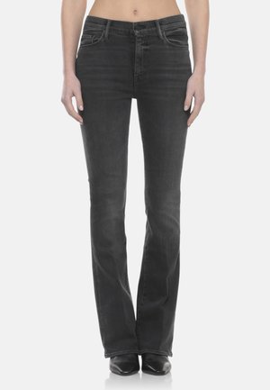 Donna che indossa jeans neri a zampa d'elefante e scarpe nere con punta appuntita, in piedi in posizione eretta con le mani rilassate ai lati.