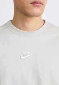 Tricou din bumbac gri deschis, cu guler rotund, având un logo Nike alb brodat pe piept. Textură netedă, cu un design minim.