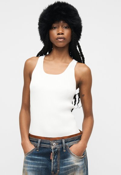 Γυναίκα με μακριά πλεξούδα, φοράει μαύρο γούνινο καπέλο, λευκό αμάνικο τοπ και ξεθωριασμένα μπλε τζιν, με τα χέρια στις τσέπες.