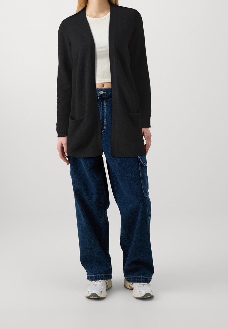 Svart cardigan med öppen front, långa ärmar och två framfickor. Bärs över en vit crop top och kombineras med lösa blå jeans och sneakers.