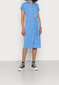 Blaues Hemdkleid mit kurzen aufgerollten Ärmeln, zwei Brusttaschen und gebundenem Taillenband. Kombiniert mit schwarzen und weißen High-Top-Sneakern.