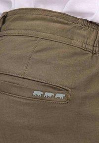 Un pantalon en coton marron présente une poche arrière ornée de trois motifs de vache gris en relief, mettant en valeur un tissu texturé et des détails de couture.