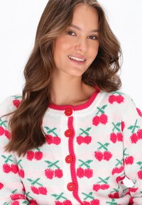 Biele pletené sako s ružovými vzormi čerešní, zelenými listami a červenými gombíkmi. Okrúhly výstrih s ružovým lemovaním. Mäkka textúra.