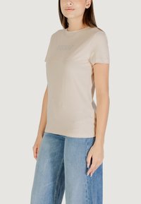 Maglietta in cotone beige a maniche corte, con un delicato logo "GUESS" in rilievo sul davanti. Abbinata a jeans a gamba larga blu chiaro.