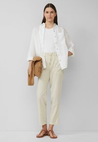 Witte linnen overhemd met knopen over een witte tanktop, gecombineerd met beige linnen broek. Het model houdt een geweven tas vast en draagt bruine sandalen.