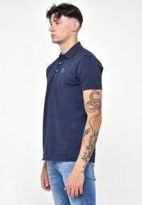 Námořnickomodrá polo tričko vyrobené z bavlny, s klasickým límcem, knoflíkovou légou s modrými knoflíky a malým logem na hrudi.