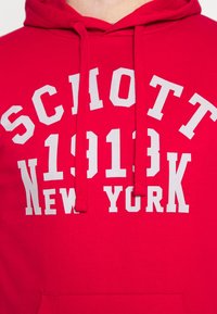 Felpa con cappuccio rossa con testo stampato bianco: "SCHOTT 1913 NEW YORK." Il materiale sembra morbido, con un design classico a girocollo e una tasca frontale.