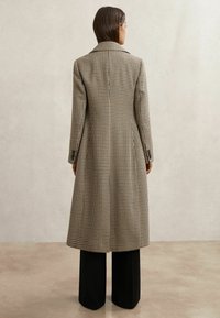 Žena s rovnými hnedými vlasmi nosí dlhý, béžovo-hnedý kabát s kockovaným vzorom a čierne široké nohavice, stojaca na lesklej podlahe.