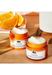 Kaksi oranssia Origins GinZing -kosteusvoidetta, joiden sisällä näkyy valkoista voidetta. Taustalla on appelsiinin viipaleita ja puinen pinta.