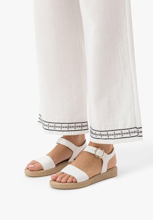 Białe sandały na płaskim obcasie z paskami wokół kostki i złotymi klamrami, noszone z białymi spodniami o szerokich nogawkach z czarnymi haftowanymi brzegami.