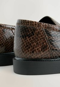 Vaaleanruskeat käärmeennahkaiset loafers-kengät kiiltävällä pinnalla, joissa on tekstuuripinta ja musta kumipohja. Suunnittelu sisältää yksityiskohtaista ompelua.