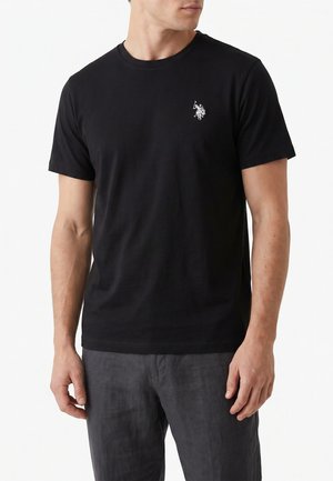 Uomo che indossa una t-shirt nera a maniche corte con un piccolo logo bianco di un giocatore di polo sul petto e pantaloni grigio scuro.