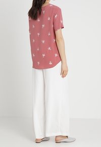 Kortærmet, lyserød top med blomstermønster lavet af letvægtigt stof, parret med brede hvide bukser og grå slip-on sko.
