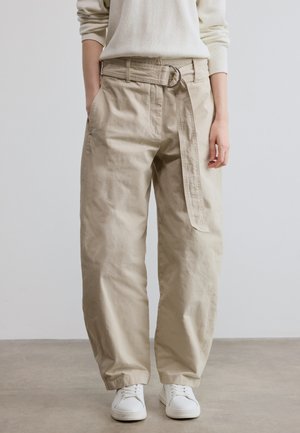 Beige Baumwollhosen mit weitem Bein, ausgestattet mit einem auffälligen Gürtel mit runder Schnalle und Seitentaschen. Tragen Sie sie mit weißen Sneakern.