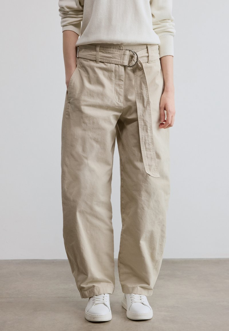 Μπεζ βαμβακερό παντελόνι με φαρδιά γραμμή, με έντονη ζώνη με κυκλικό αγκράφα και πλευρικές τσέπες. Φορεμένο με λευκά αθλητικά παπούτσια.