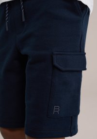 Un short en coton bleu marine présente une poche extérieure sur le côté, avec des détails de couture et une étiquette logo sur le bas.