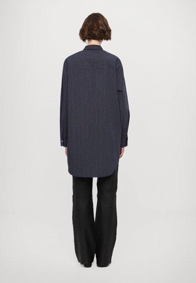 Camisa de manga larga azul oscuro con rayas verticales rojas. La vista posterior muestra un dobladillo curvado y detalles de botones en los puños. Combinada con pantalones negros de pierna ancha.