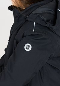 Schwarze, wasserdichte Jacke mit Kapuze und reflektierenden Akzenten. Sie verfügt über ein kreisförmiges Logo auf der linken Schulter und eine glatte Stoffstruktur.