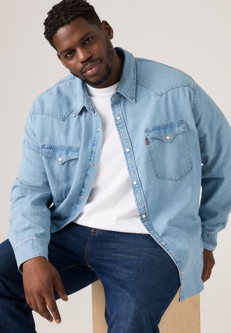 Light blue denim shirt with long sleeves, two chest pockets, and a button-down front, worn over a white shirt. Classic design, relaxed fit.