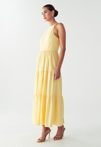 Nainen seisoo keltaisessa olkaimettomassa, kerroksellisessa maksimaksissa, jossa on sivuleikkaukset, päällään kullanväriset renkaat ja nudeväriset, korsettimaiset korkokengät.