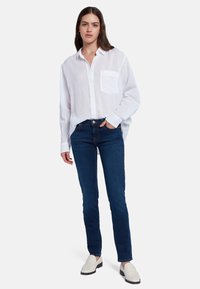 Chemise blanche à manches longues avec un poche poitrine, associée à un jean ajusté bleu foncé et des mocassins blancs. Design simple et épuré.