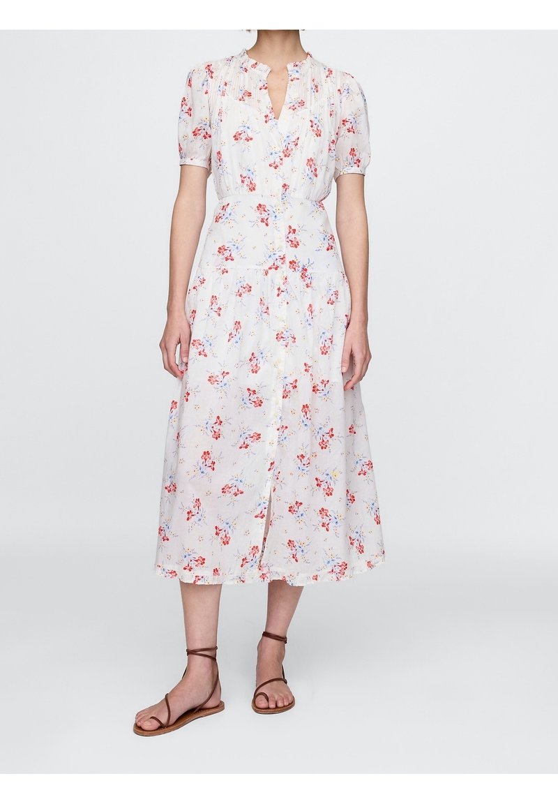 Femme portant une robe midi blanche avec un imprimé floral rouge et bleu, manches courtes bouffantes, boutons à l'avant et sandales plates marron à bride cheville.