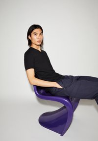 Zwart V-hals T-shirt gedragen met marineblauwe broek, casual zittend op een gebogen paarse kunststof stoel tegen een lichtgrijze achtergrond.