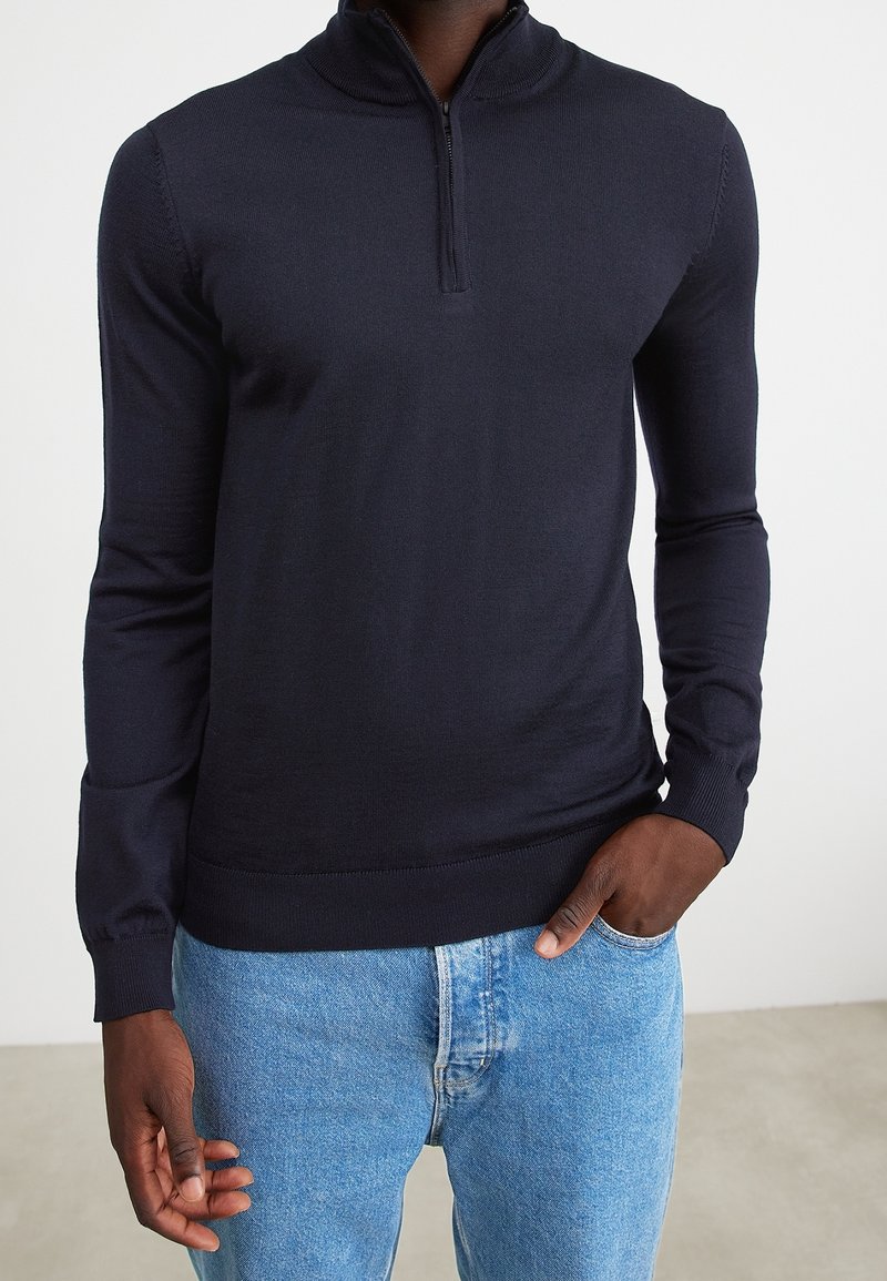 Homme portant un pull bleu marine ajusté à zip court avec manches longues et un jean bleu clair, main dans la poche, sur fond blanc uni.