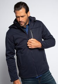 Marinefarbene Softshelljacke mit Kapuze, ausgestattet mit einem Reißverschluss vorne, Seitentaschen und Nähdetails. Getragen über einem blau karrierten Hemd.