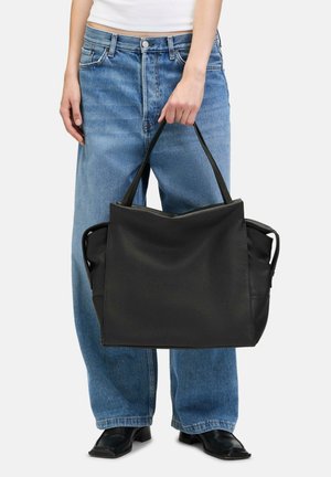 Sac en cuir noir avec une texture lisse, de forme rectangulaire, deux poignées supérieures et un intérieur spacieux. Associé à un jean large bleu clair.