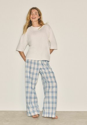 Smiling woman stands barefoot against beige background, wearing loose white short-sleeve top and blue-and-white checkered wide-leg pants.