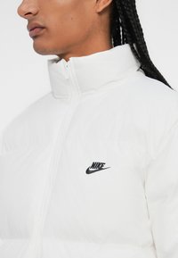 Bijela puffer jakna s visokim ovratnikom, s crnim Nike logom na prsima, izrađena od sjajnog, podstavljenog materijala.