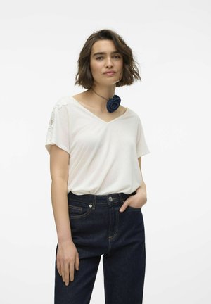 Hvid, kortærmet V-hals top med blonderdetaljer på ærmerne, parret med mørkeblå jeans. En denimrose-accessorie bæres om halsen.