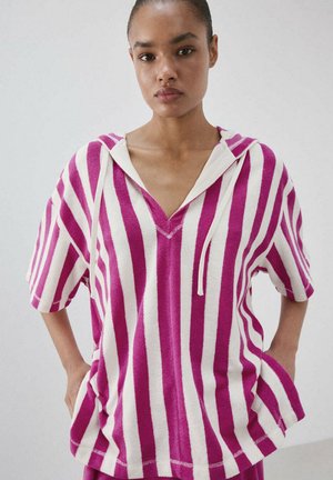 Mujer con una sudadera con capucha de manga corta con rayas verticales rosas y blancas, de pie con las manos en los bolsillos contra un fondo liso.