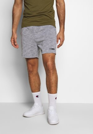 Homme portant un short décontracté gris, un T-shirt vert olive, des chaussettes blanches Champion et des baskets blanches, debout devant un fond blanc uni.