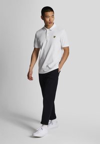 Polo bianco con logo giallo sul petto, abbinato a pantaloni scuri e sneakers bianche. Tessuto liscio, design con colletto, vestibilità classica.