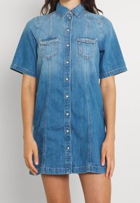 Vestido de camisa de mezclilla en azul claro, con mangas cortas, cierre de botones en el frente, dos bolsillos en el pecho y detalles bordados a lo largo de los hombros.