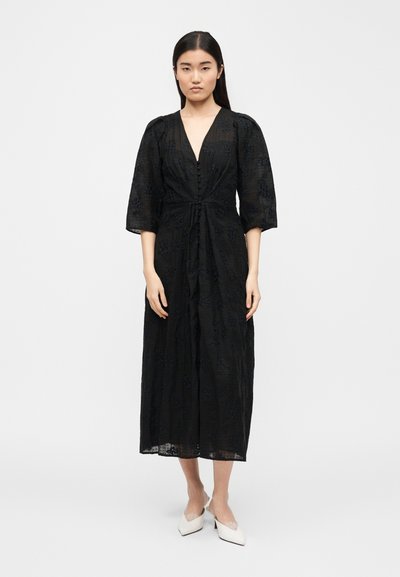 Νεαρή γυναίκα στέκεται φορώντας μαύρο midi φόρεμα με υφή, 3/4 μανίκια και λευκές μυτερές χαμηλές γόβες, μπροστά σε απλό ανοιχτό φόντο.