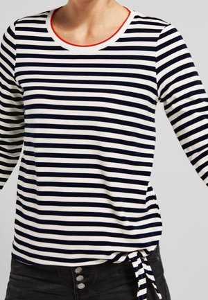 Person trägt ein langärmeliges Shirt mit schwarzen und weißen horizontalen Streifen, rotem Halsausschnitt und gebundenem Detail in Taillennähe, kombiniert mit einer schwarzen Hose.